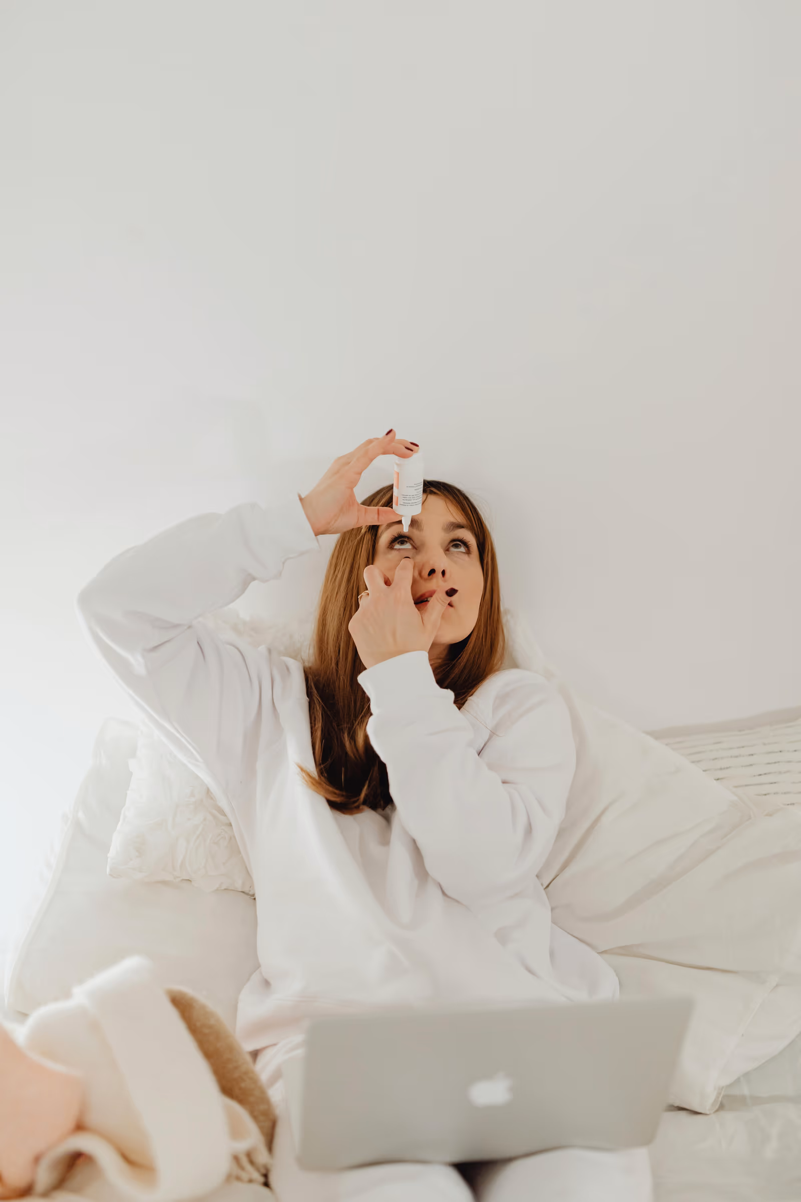 Mulher de pijama branco aplicando colírio sentada na cama com notebook, cena de auto-cuidado em casa durante o pós-operatório. Foto: Karola G / Pexels. Imagem ilustrativa de banco de imagens.